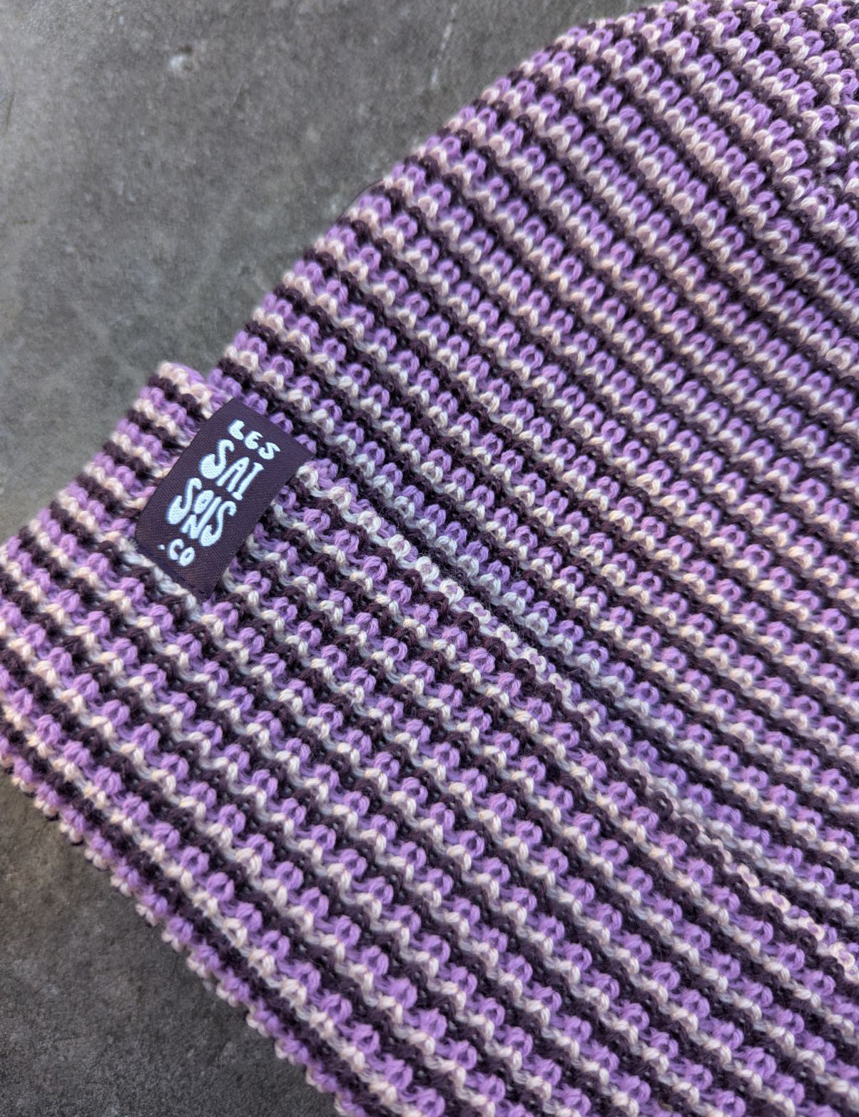 zoom on the purple beanie with a purple les Saisons tag on a gray concrete surface, made in Canada.