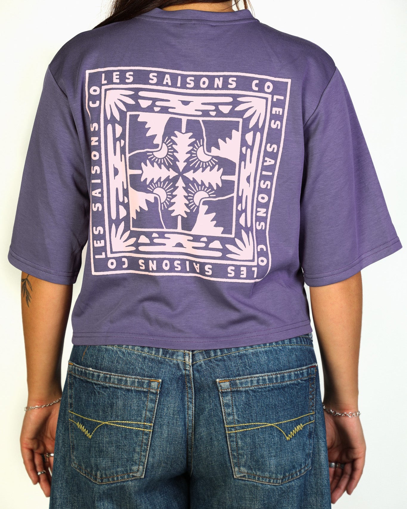A person shown from the back wearing a purple cropped T-shirt with a large white graphic print reading 'LES SAISONS CO' and blue jeans against a plain light background.