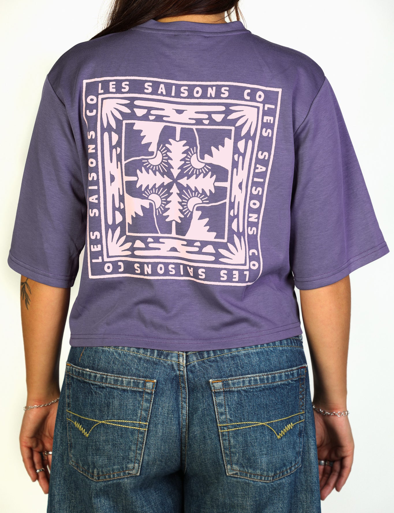 A person shown from the back wearing a purple cropped T-shirt with a large white graphic print reading 'LES SAISONS CO' and blue jeans against a plain light background.