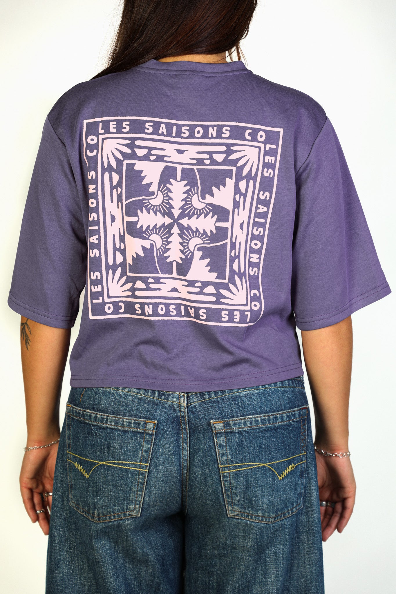 A person shown from the back wearing a purple cropped T-shirt with a large white graphic print reading 'LES SAISONS CO' and blue jeans against a plain light background.