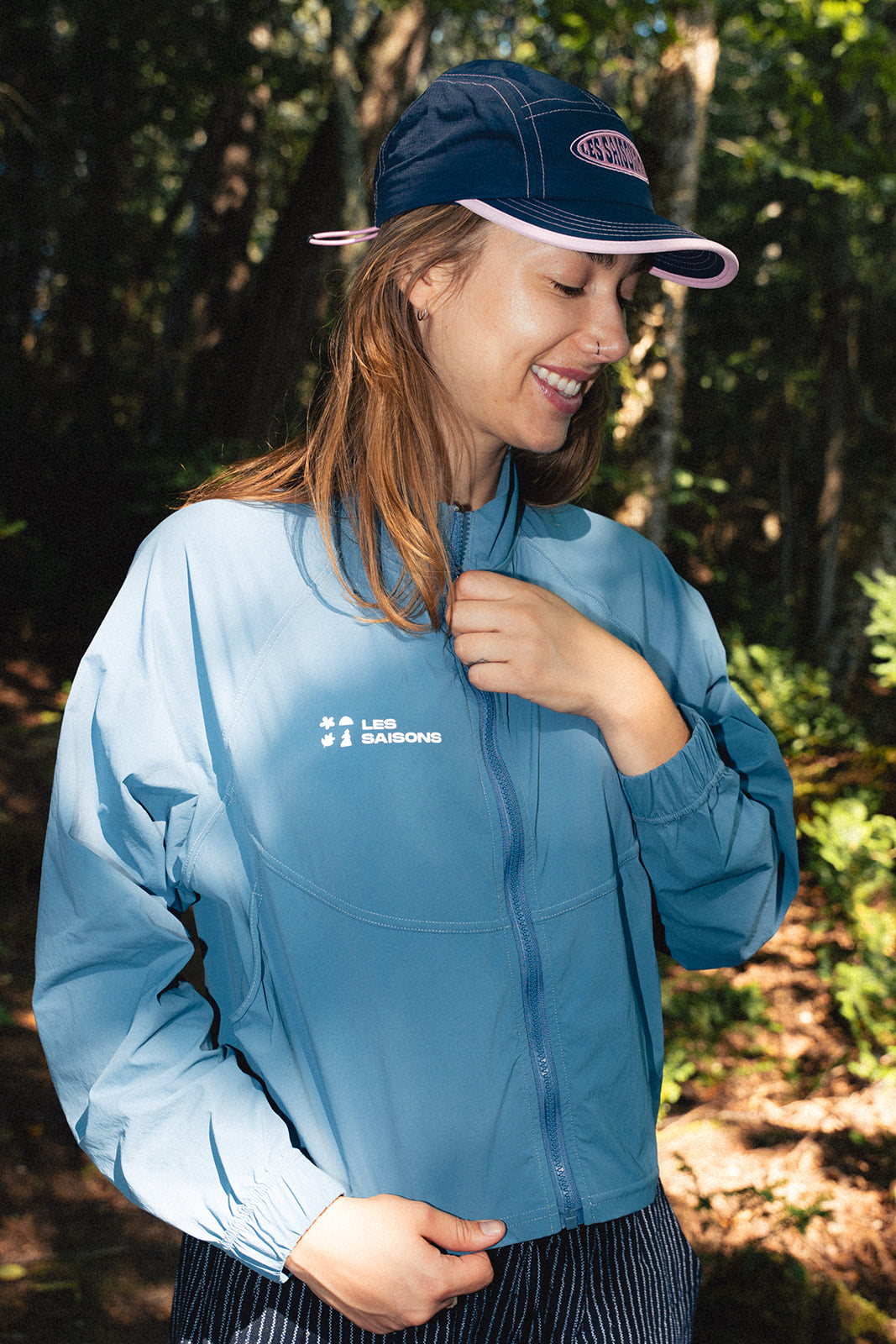Woman wearing a blue active rain jacket from les Saisons and cap outdoors with trees in the background