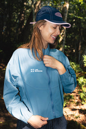 Woman wearing a blue active rain jacket from les Saisons and cap outdoors with trees in the background