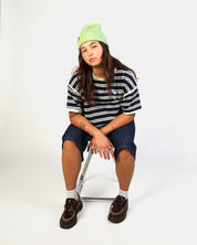 A model sitting on a metal folding chair wearing a lime green beanie, a navy and white striped t-shirt, denim shorts, white socks, and dark brown lace-up shoes against a plain white studio background.