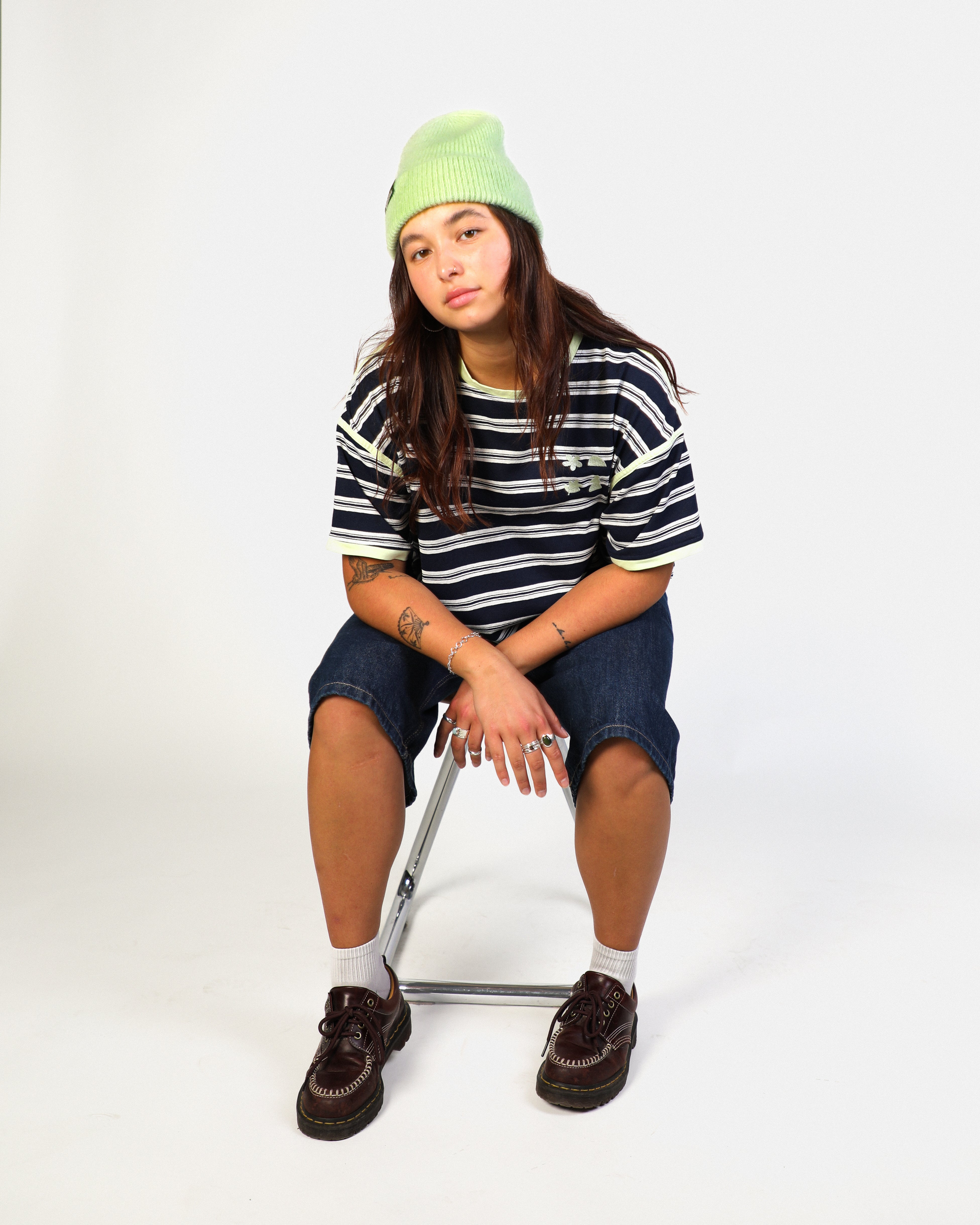 A model sitting on a metal folding chair wearing a lime green beanie, a navy and white striped t-shirt, denim shorts, white socks, and dark brown lace-up shoes against a plain white studio background.