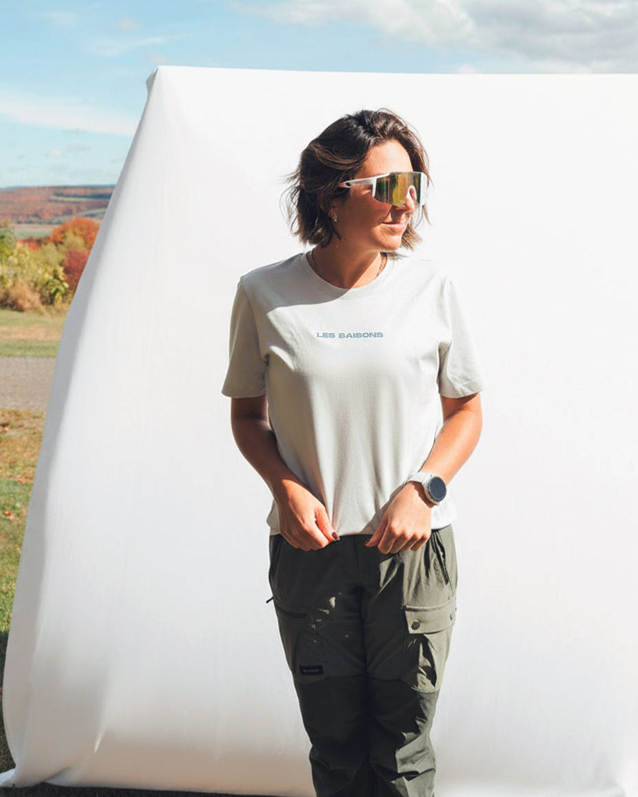 Person standing outdoors with a large white background wearing sunglasses and a grey les Saisons t-shirt.